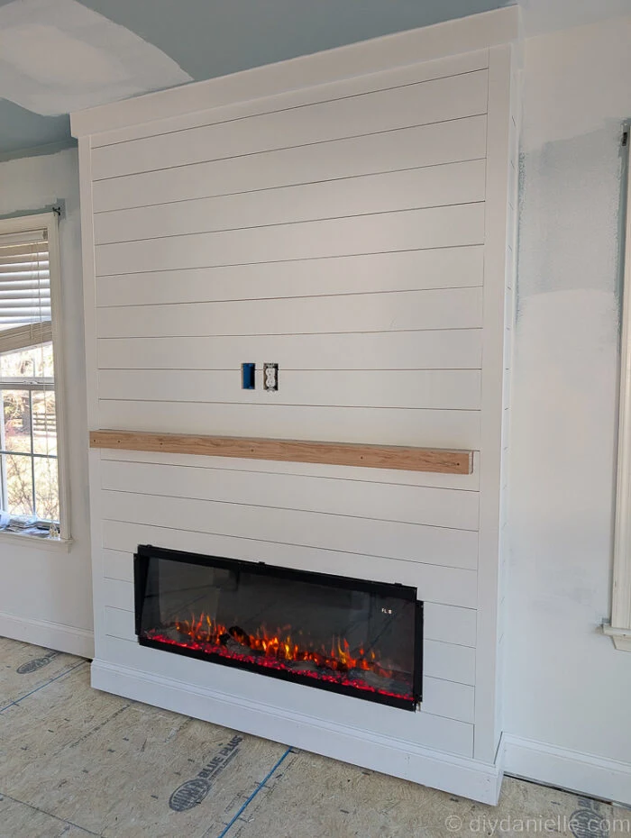 shiplap fireplace surround for an electric fireplace with trim, fireplace turned on, and a small mantle. There's outlets for a tv above.