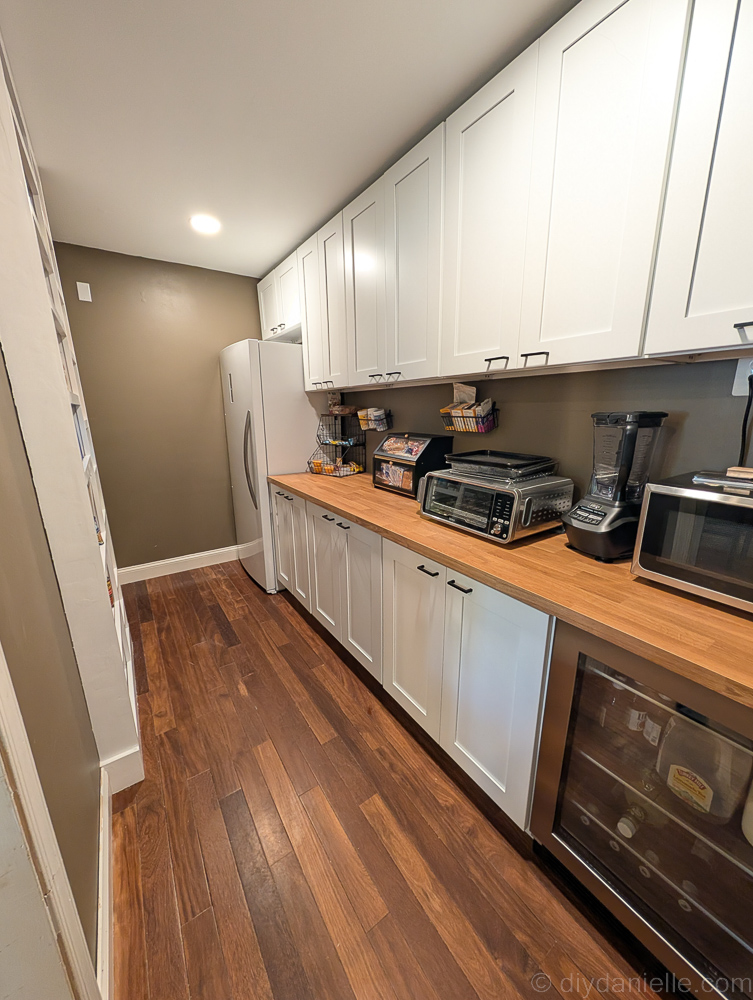 DIY Pantry made by dividing the small dining room into two rooms: the pantry and a mudroom. The pantry has white shelving for cans (4" deep) and the opposite side has a large freezer, a small mini fridge for drinks, and white cabinets. The countertop is a natural wood color (IKEA countertop).