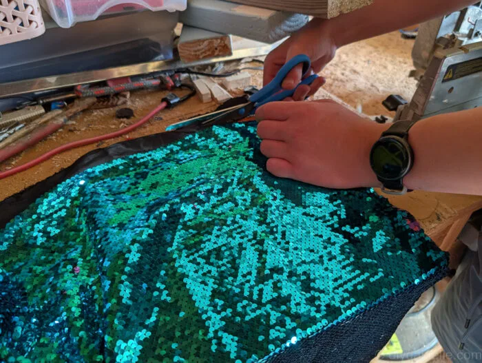 Sequin fabric being trimmed with scissors to match the size of a wooden board for a DIY fidget project.