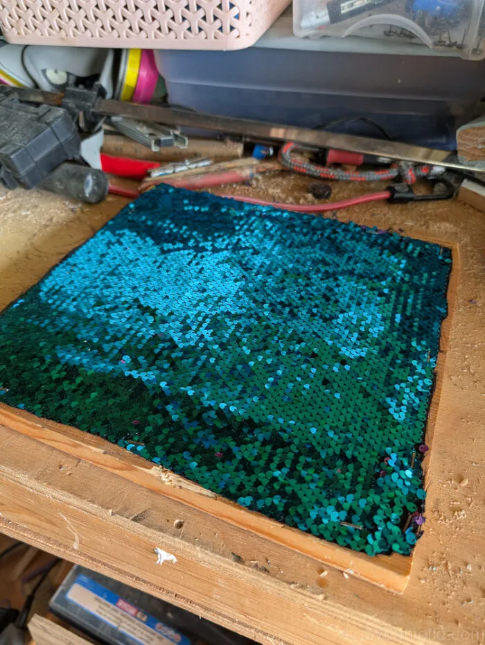 Hands using a stapler to attach sequin fabric to the top of a wooden board for a DIY fidget board project.
