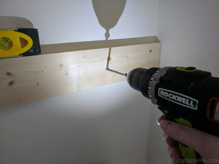 DIY over-the-toilet shelf being mounted, showing screws aligned with studs and the board leveled for proper installation.