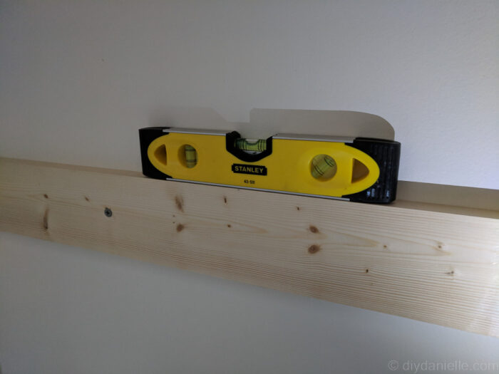Level board being hung above a toilet for DIY over-the-toilet storage, with screws placed in studs for secure installation.