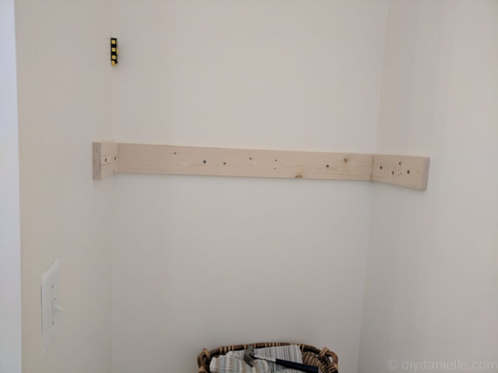 DIY over-the-toilet shelf being installed, showing proper leveling and use of studs or drywall anchors for secure mounting.