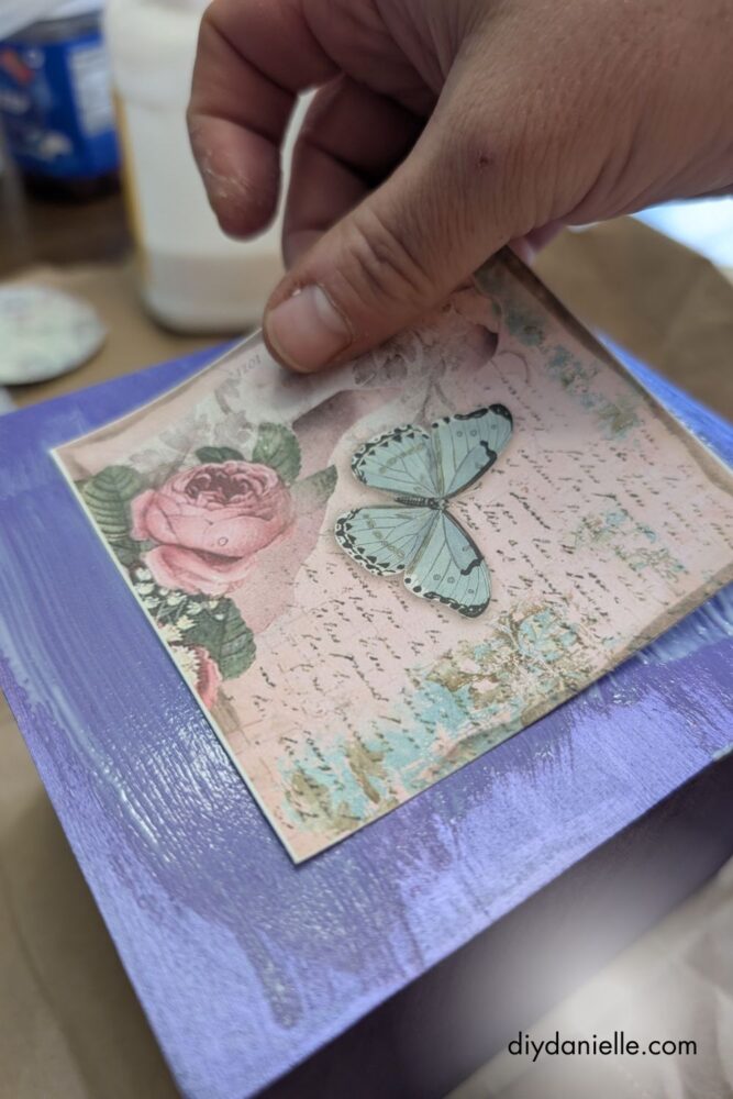 Pressing scrapbook paper onto a tissue box with mod podge, smoothing out air bubbles.