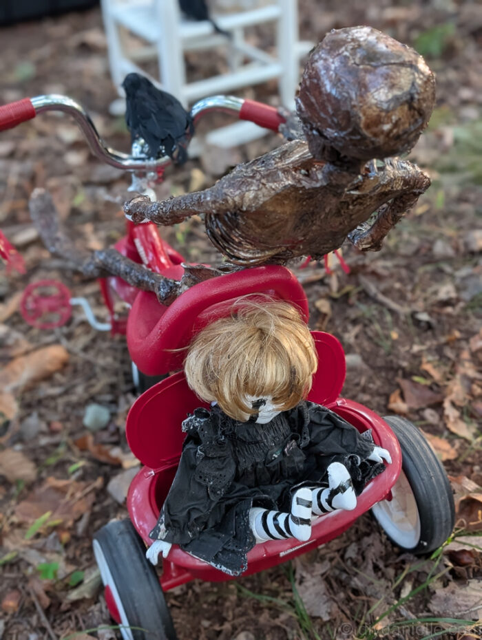 Skeleton positioned on a tricycle with a baby doll placed in the back cubby as part of a spooky Halloween scene.