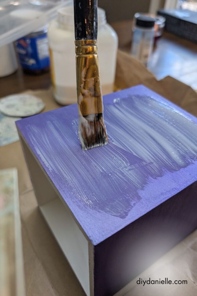 Applying a thin layer of mod podge to a painted tissue box to attach decorative scrapbook paper.