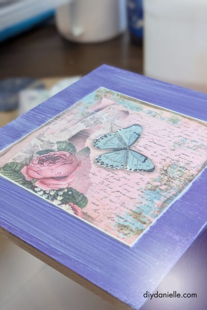 Applying a top layer of mod podge over scrapbook paper on a tissue box, smoothing out air bubbles.