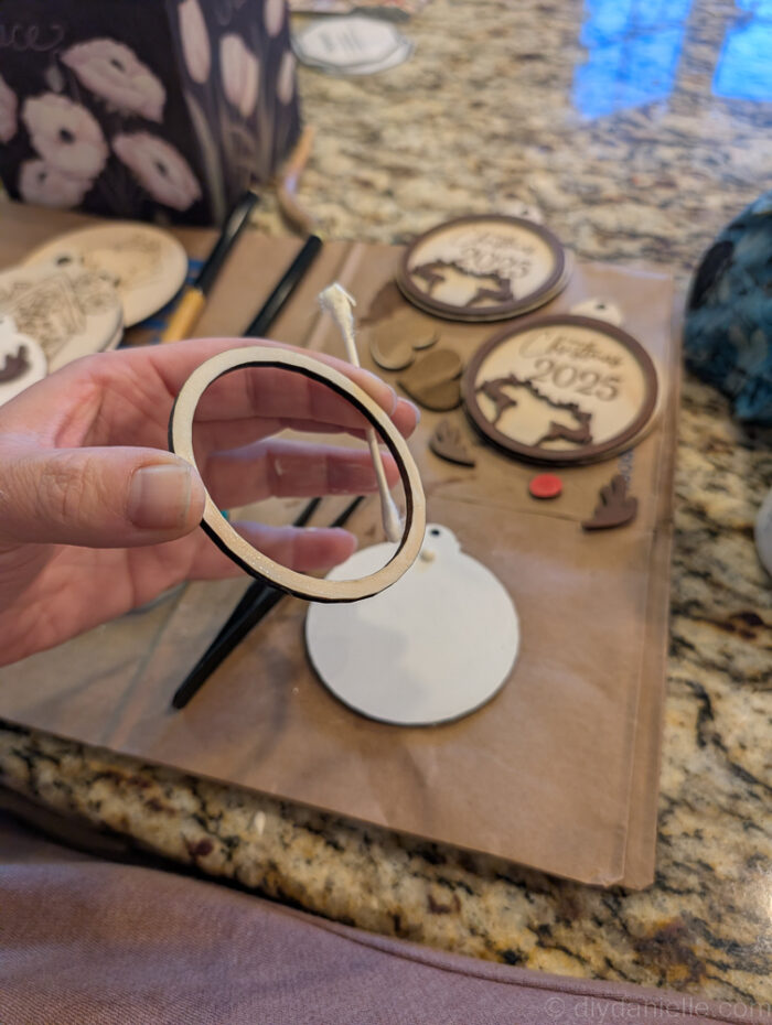 Hands using a Q-tip to apply wood glue to the back of a small ornament piece, assembling a two-layer wooden Rudolph ornament with tweezers for tiny details.