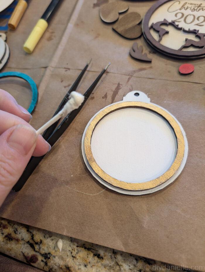 Hands using a Q-tip to apply wood glue to the back of a small ornament piece, assembling a two-layer wooden Rudolph ornament with tweezers for tiny details.