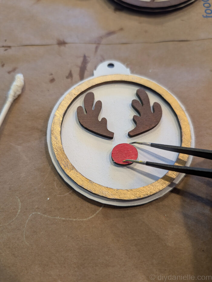 Hands using a Q-tip to apply wood glue to the back of a small ornament piece, assembling a two-layer wooden Rudolph ornament with tweezers for tiny details.