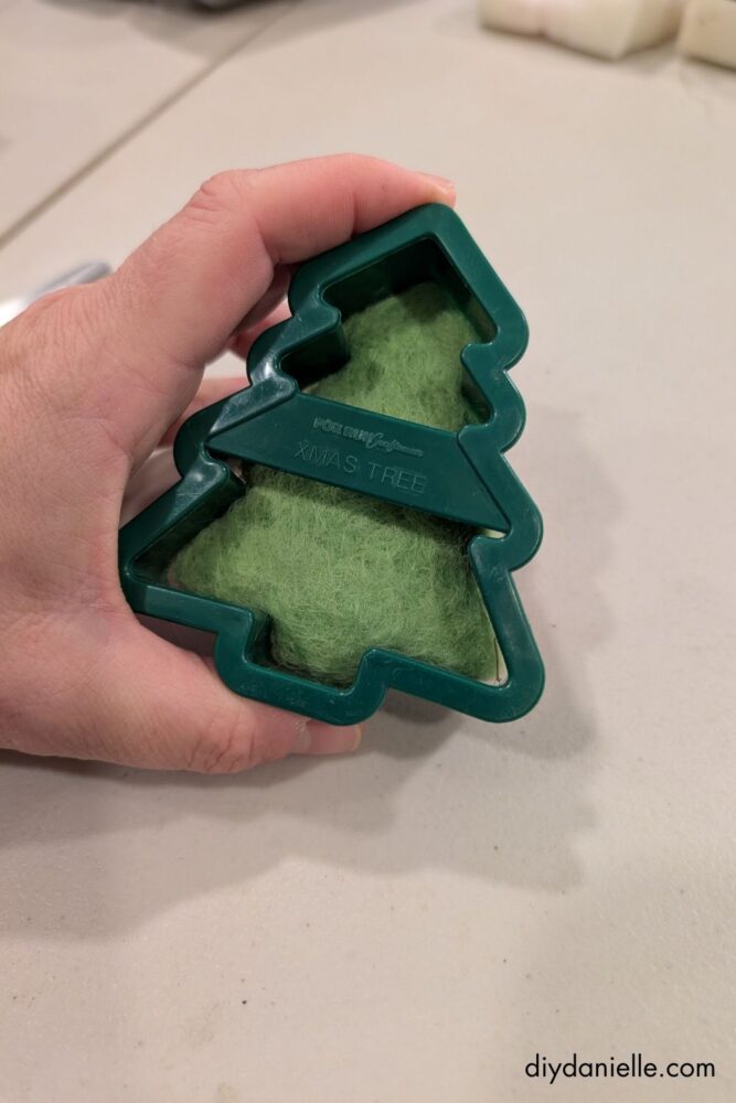 Hands placing wool roving into a cookie cutter on a felting pad to create a DIY felted ornament in holiday shapes.