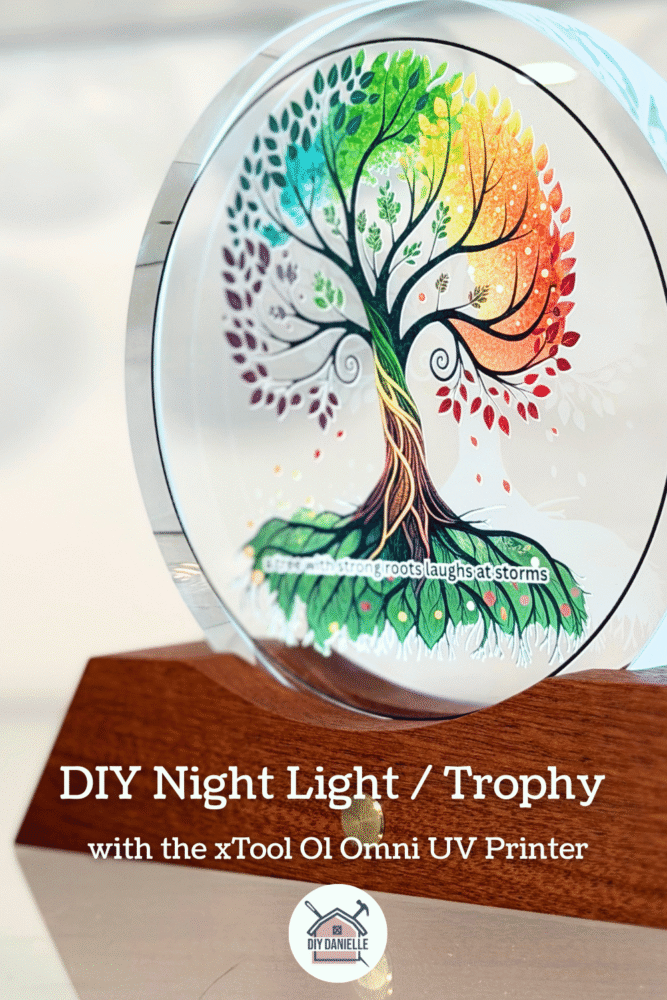This is a close up of a round, glass plaque sitting in a wood base with a light on it. The glass has a colorful version of the Tree of Life printed on it with the words "a tree with strong roots laughs at storms" at the bottom. Text says "DIY Night Light / Trophy with the xTool O1 Omni UV Printer"
